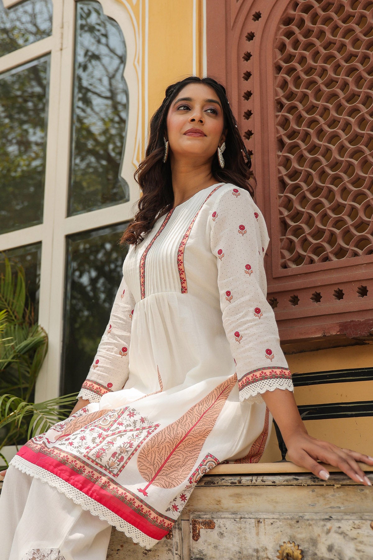 Woman in a white traditional outfit with red and pink embroidery sitting on a window ledge.