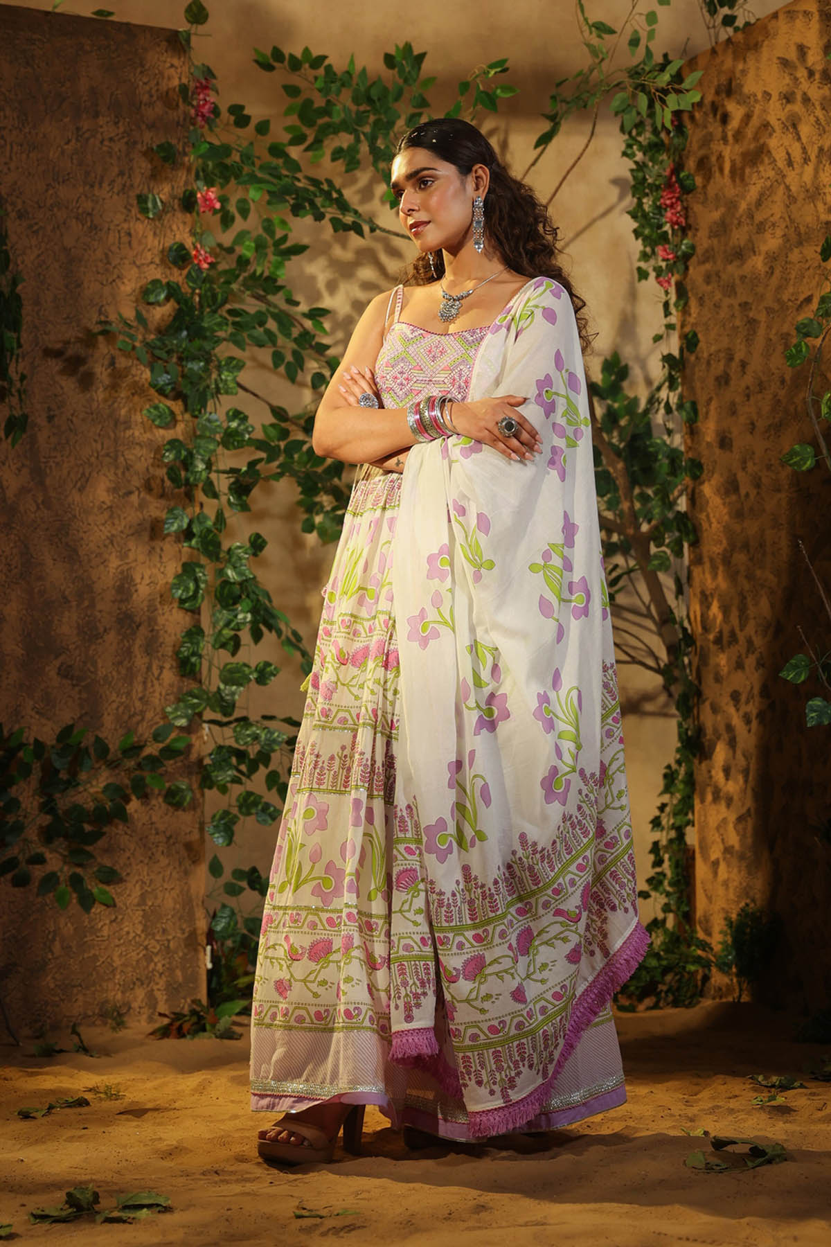 Woman in a white saree with floral patterns standing against a textured wall with greenery.