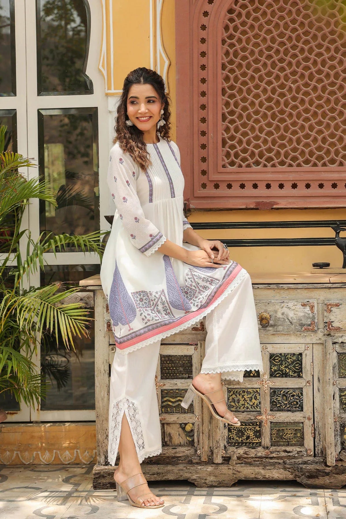 Woman in a white traditional outfit sitting on a wooden bench with a decorative background