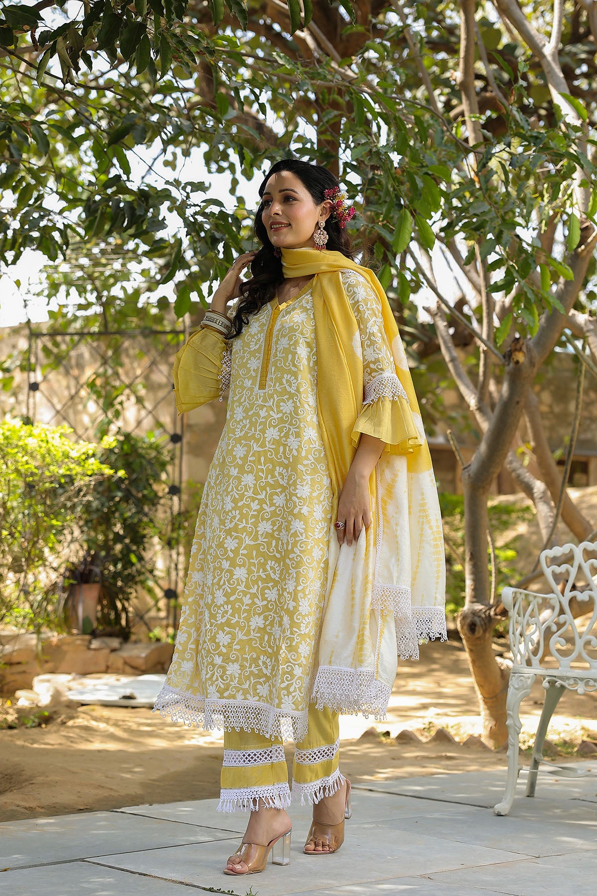 Woman in a yellow and white traditional outfit standing outdoors with trees and a camel in the background.
