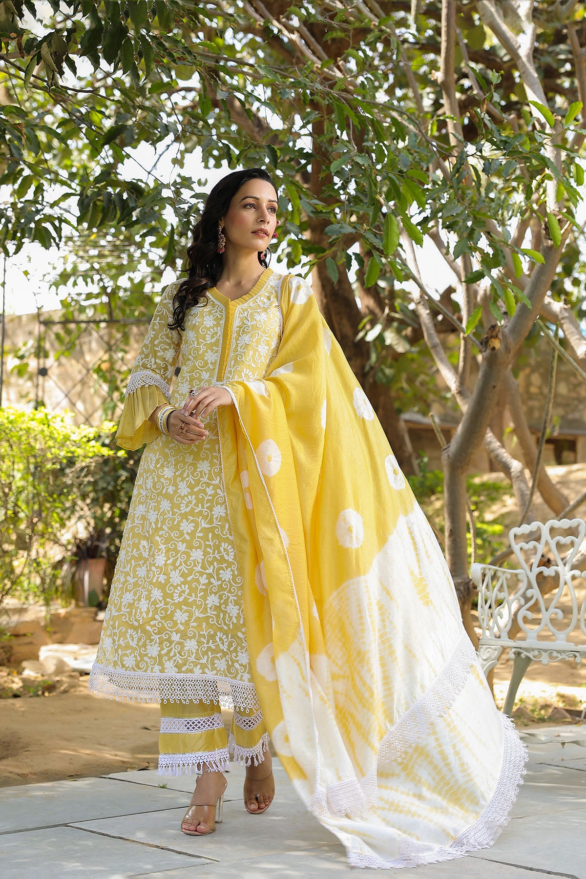 Woman in a yellow and white traditional outfit standing outdoors with greenery.