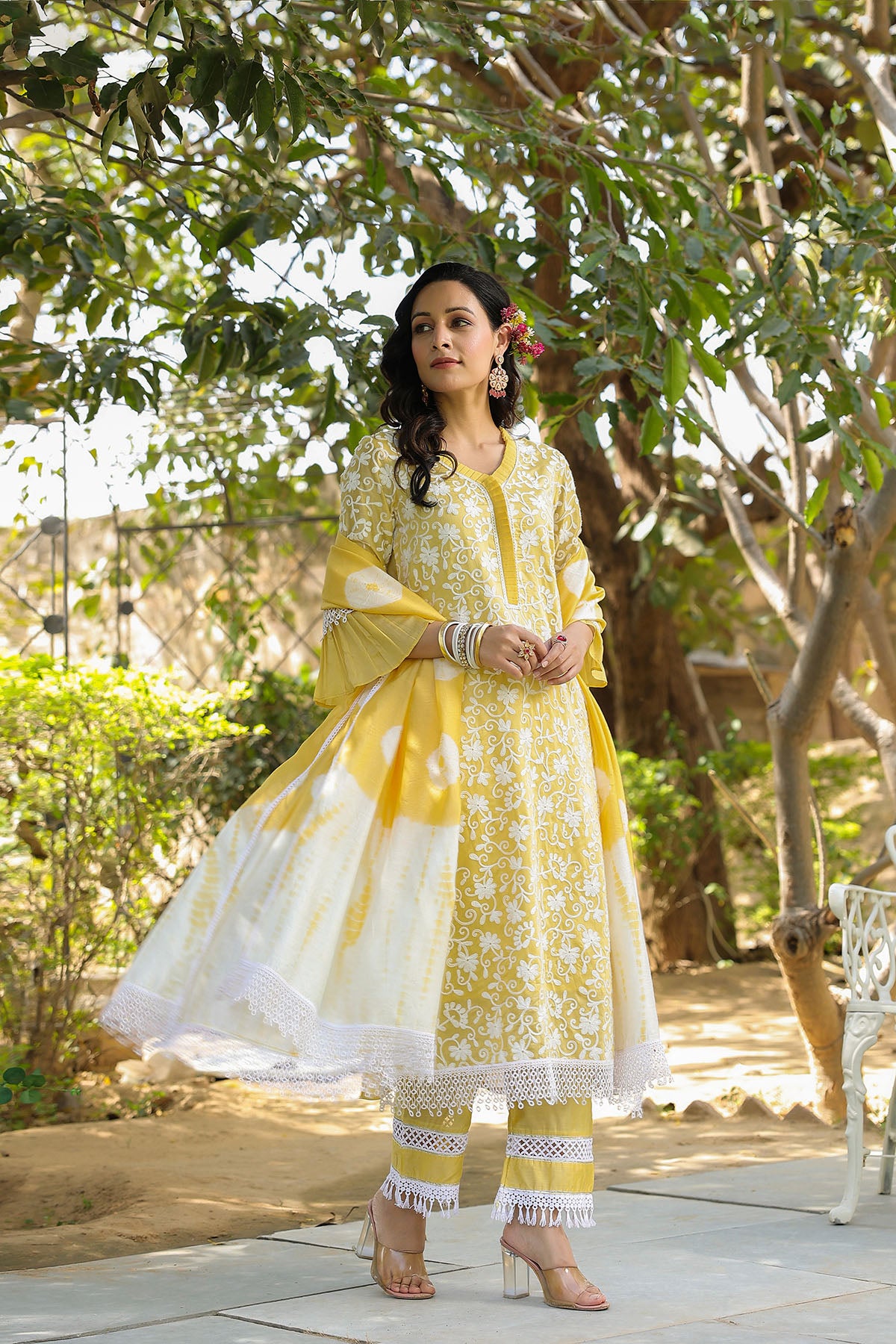 Woman in a yellow and white traditional outfit standing outdoors with greenery in the background