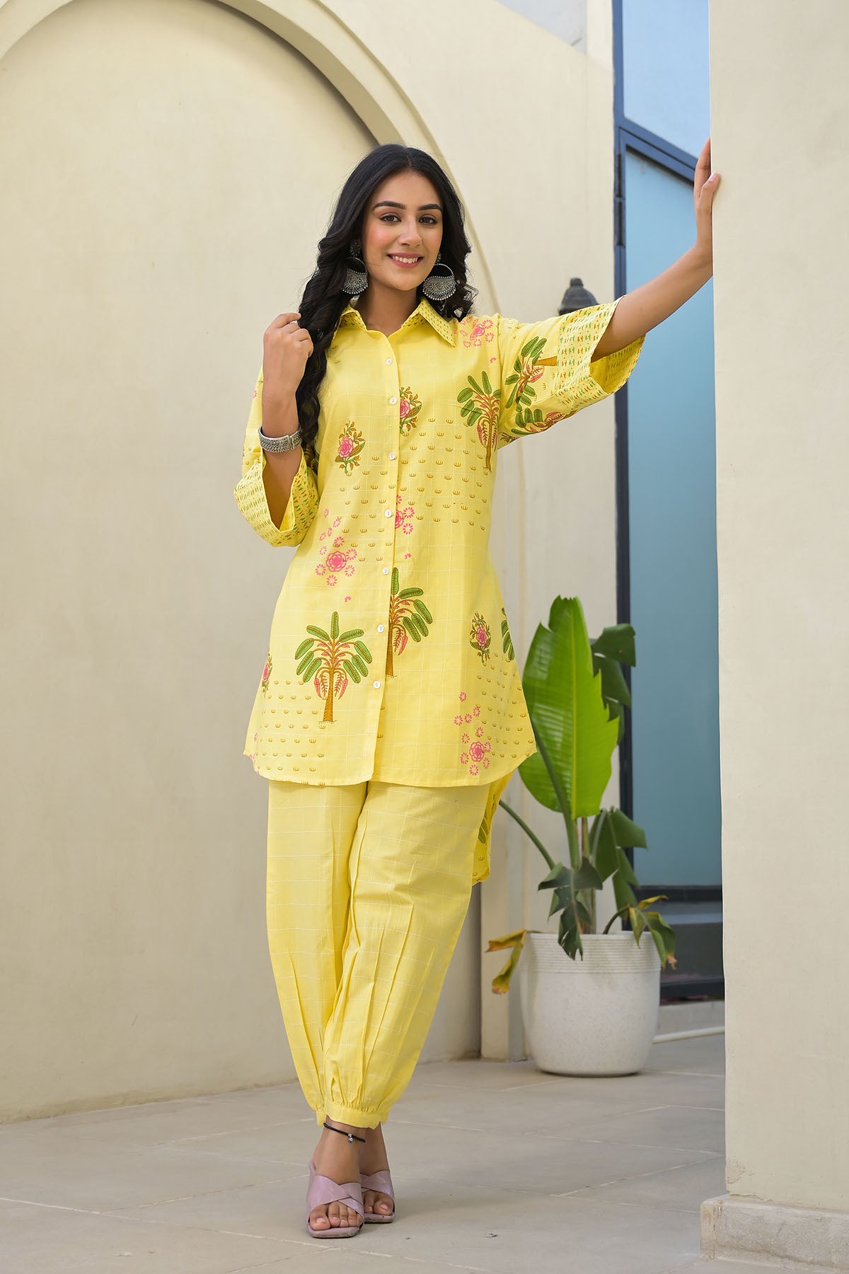 Woman wearing a yellow embroidered outfit standing indoors with a plant in the background
