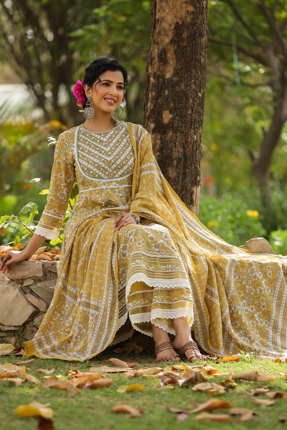 Woman in a yellow and white traditional outfit sitting outdoors with greenery around.