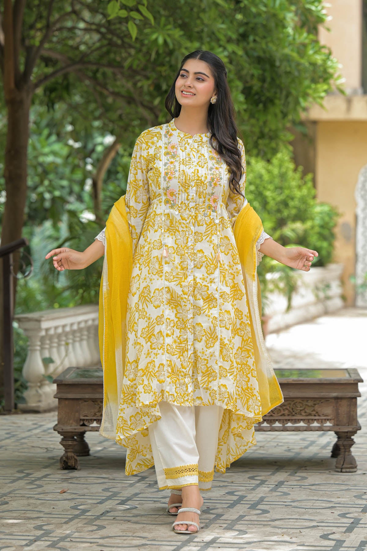 Woman in a yellow and white floral outfit standing outdoors with greenery in the background