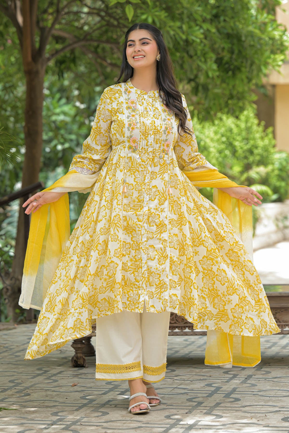 Woman in a yellow and white traditional outfit standing outdoors with greenery in the background