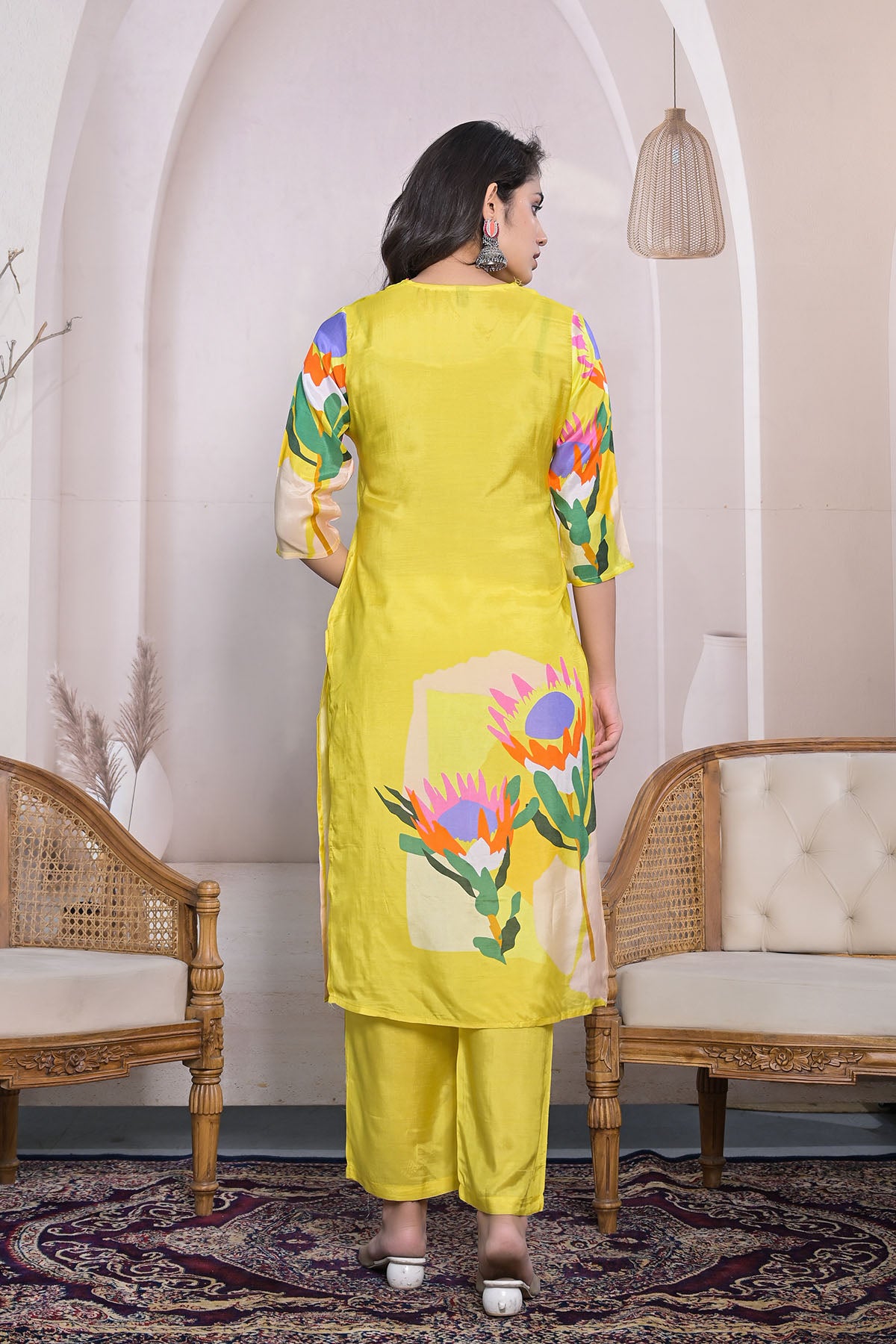 Woman wearing a yellow kurta with floral patterns in a room with wicker chairs and a decorative rug.