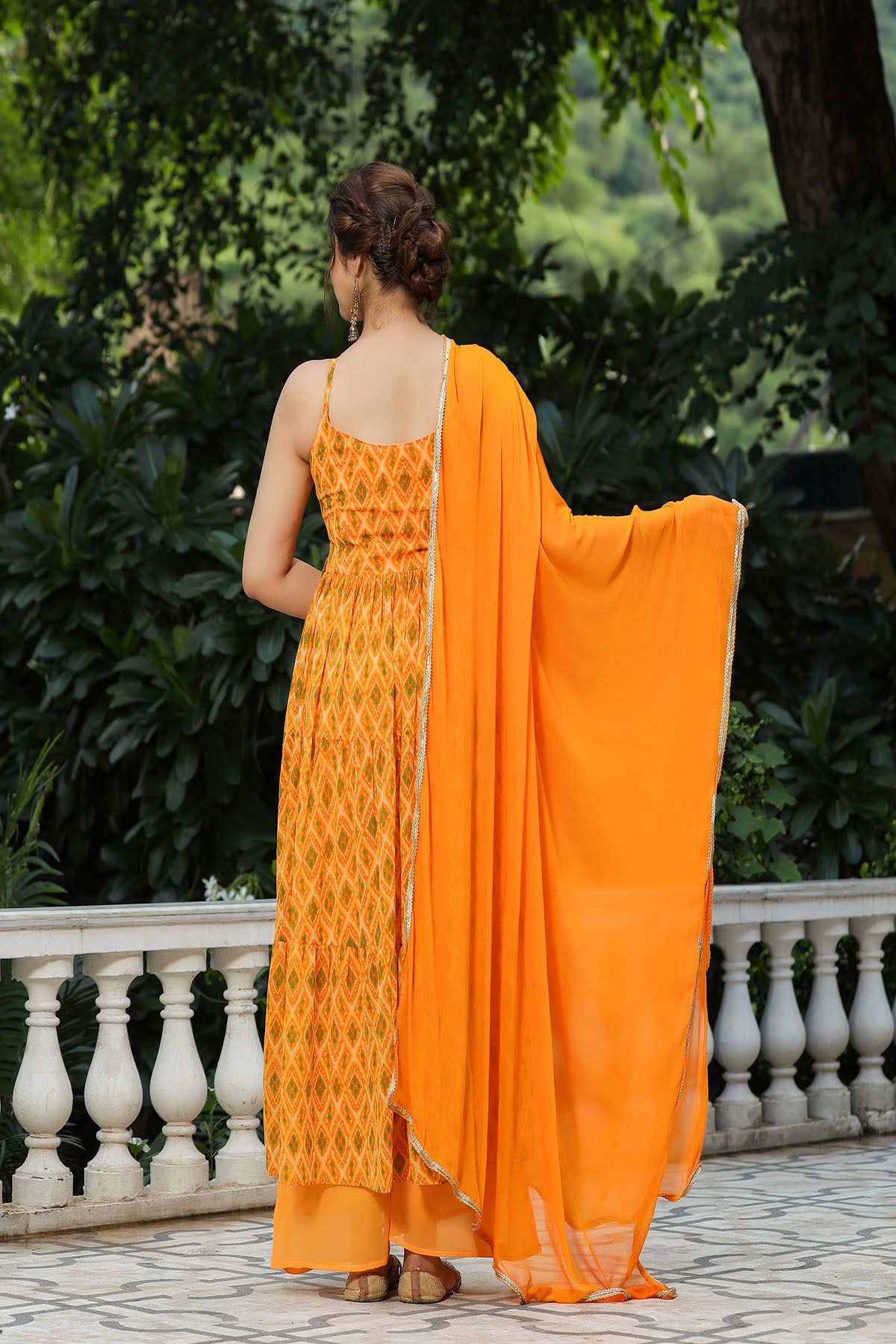 Woman in an orange saree with a patterned dress, standing outdoors with greenery in the background.