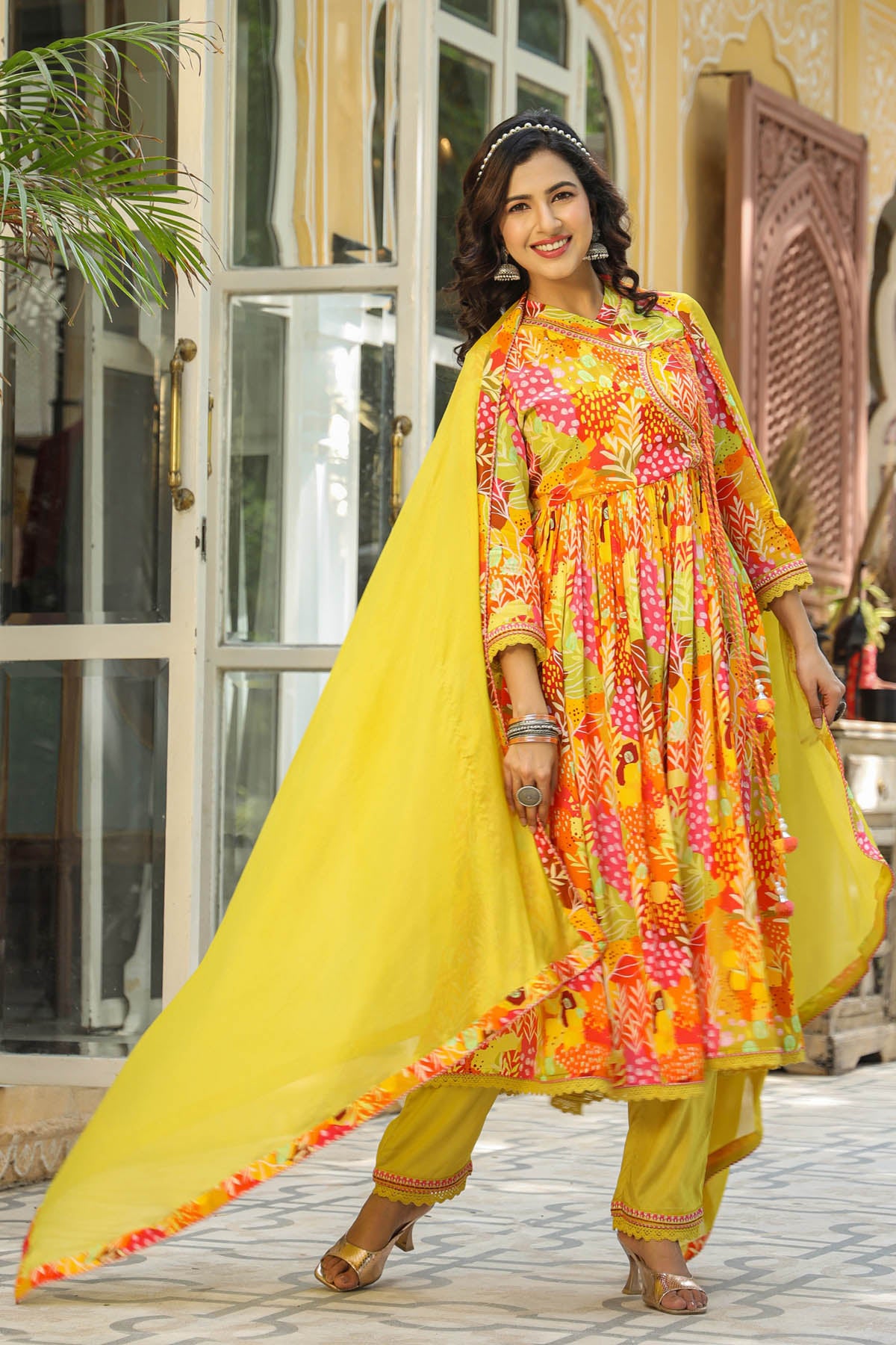 Woman in a colorful traditional outfit with a yellow dupatta standing in front of a building entrance.