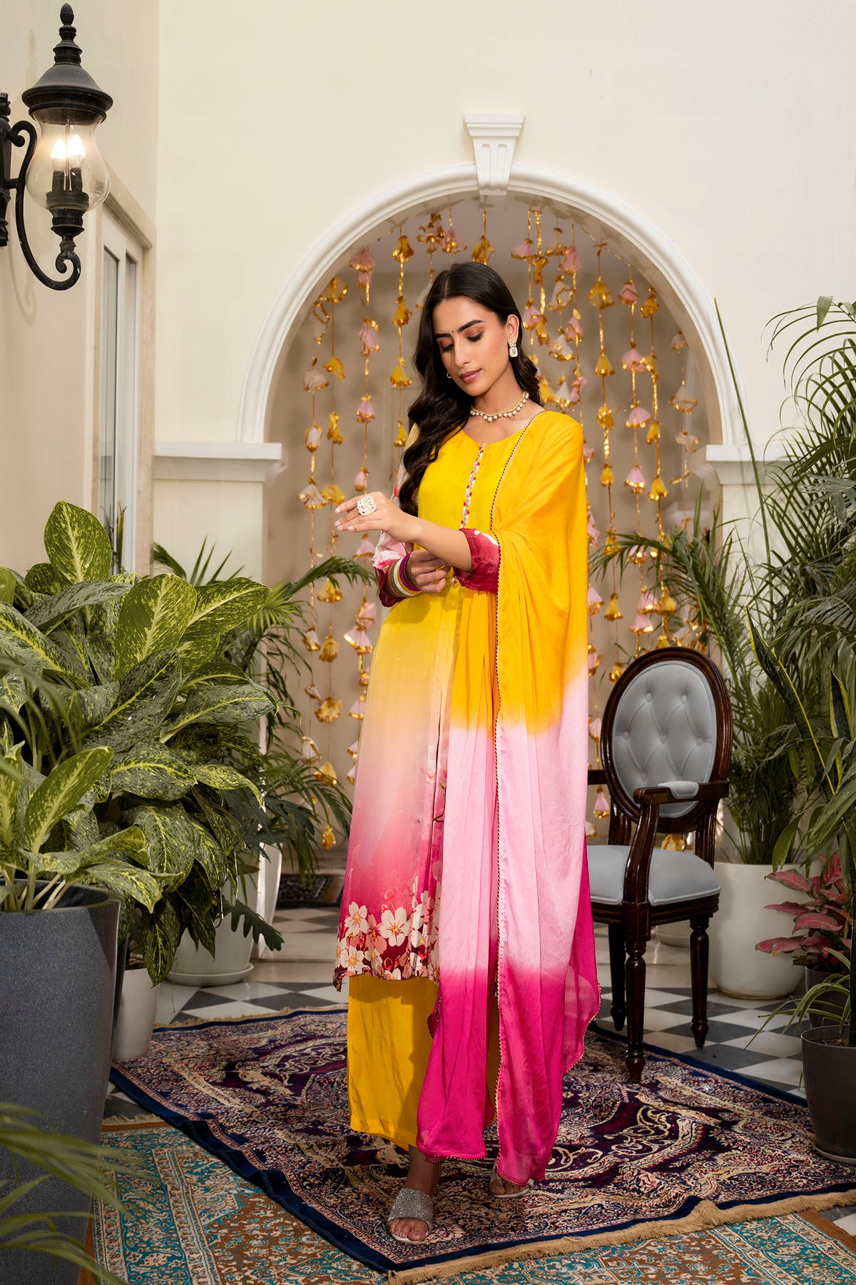 Woman in a yellow and pink gradient dress standing in an elegant indoor setting with plants and decorative archway.