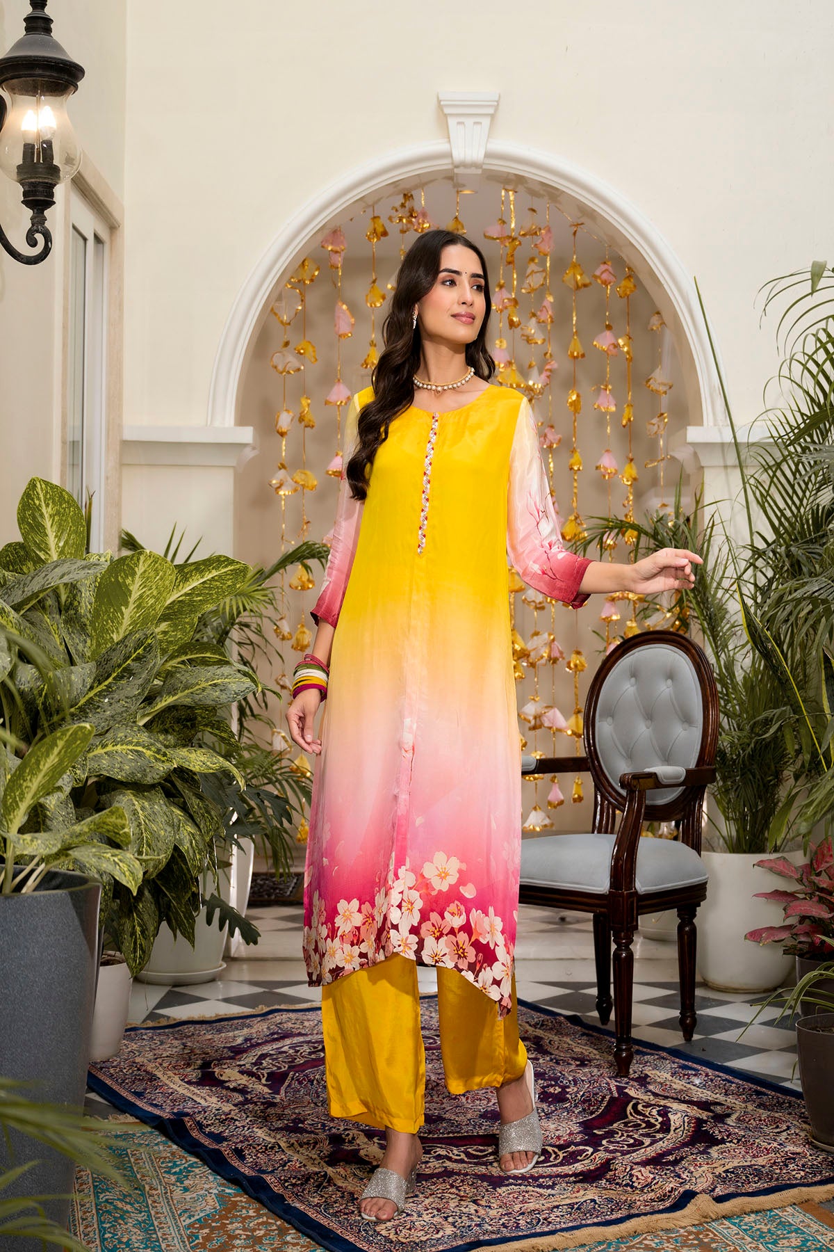 Woman in a yellow and pink dress standing in an elegant indoor setting with plants and a chair.