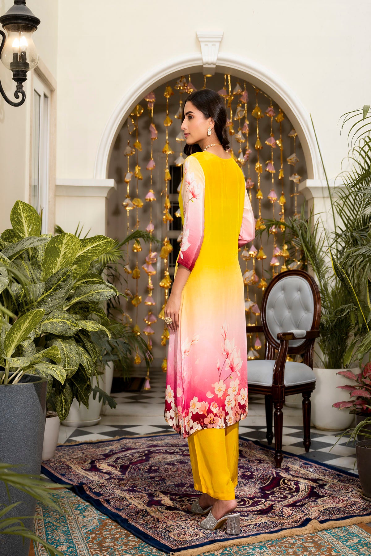 Woman in a yellow and pink dress standing in an outdoor setting with plants and a decorative wall.