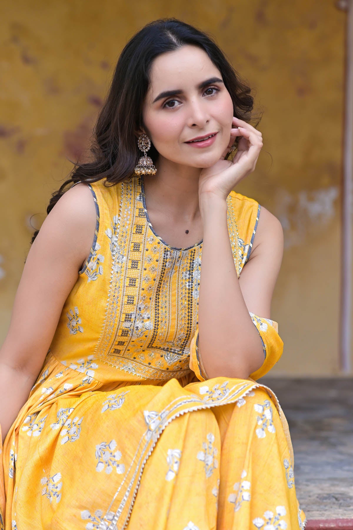Woman in a yellow traditional outfit with floral patterns, sitting against a blurred background.