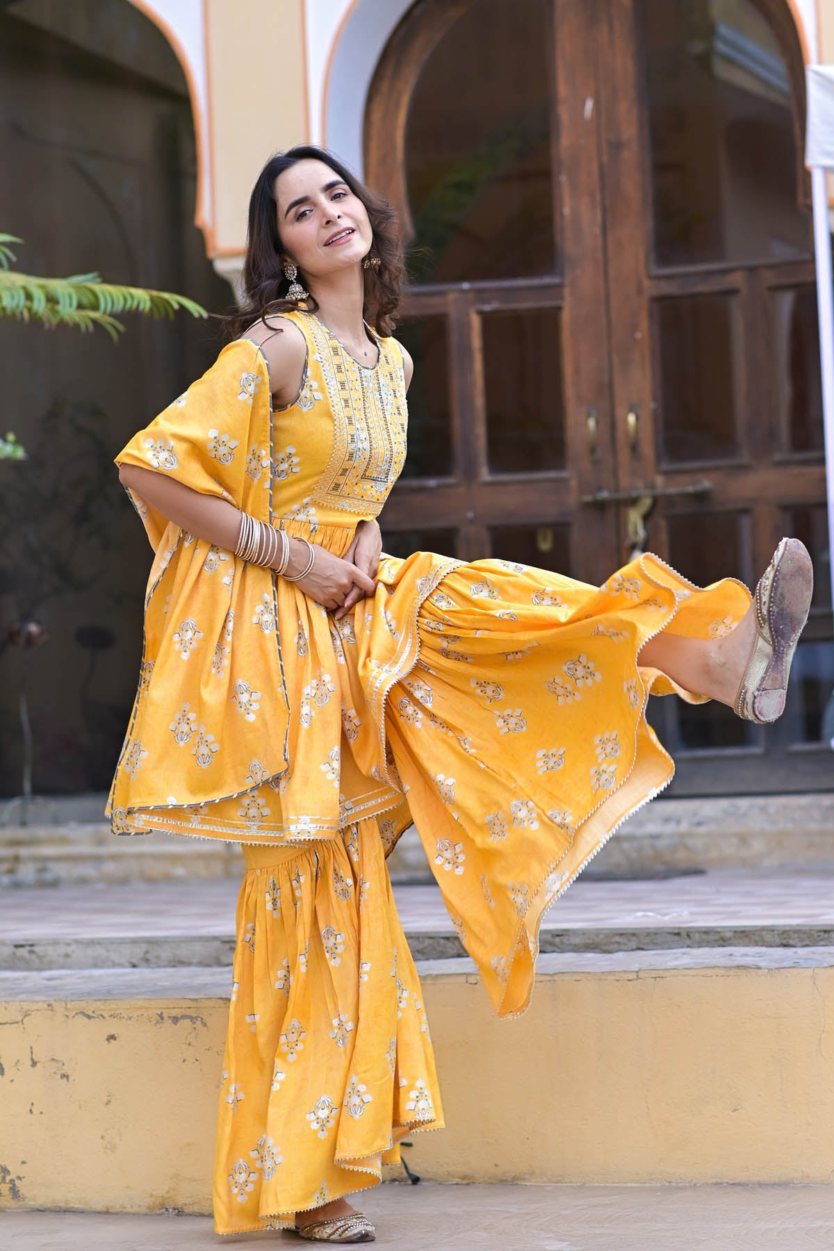 Woman in a yellow saree with a floral pattern standing on steps outdoors.